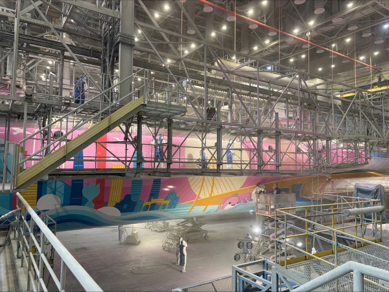 Wide view of the aircraft in a facility during the coating process, surrounded by scaffolding and with several workers throughout the frame.