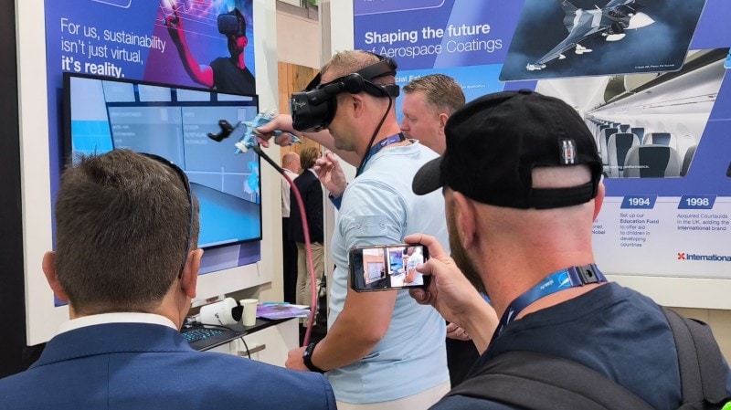 A trade show attendee wearing a VR headset and holding a spray-paint controller participates in a virtual reality paint simulation at the Aerospace Coatings booth, while other attendees observe on the monitor.