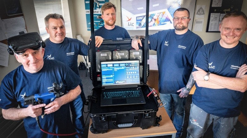 The team of technicians at IAC showcasing their VR painting equipment, standing around a table with a large electronics case containing a laptop connected to the VR headset and spraygun controller held by the trainer on the left.