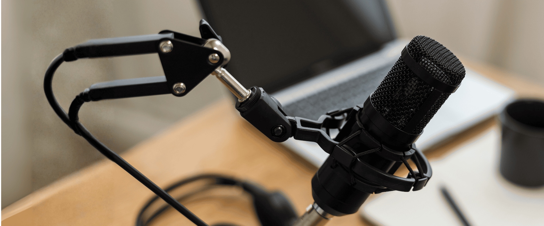 Close-up of a podcast microphone on a desk with a laptop, notebook, pen, coffee mug, and headphones in the background.