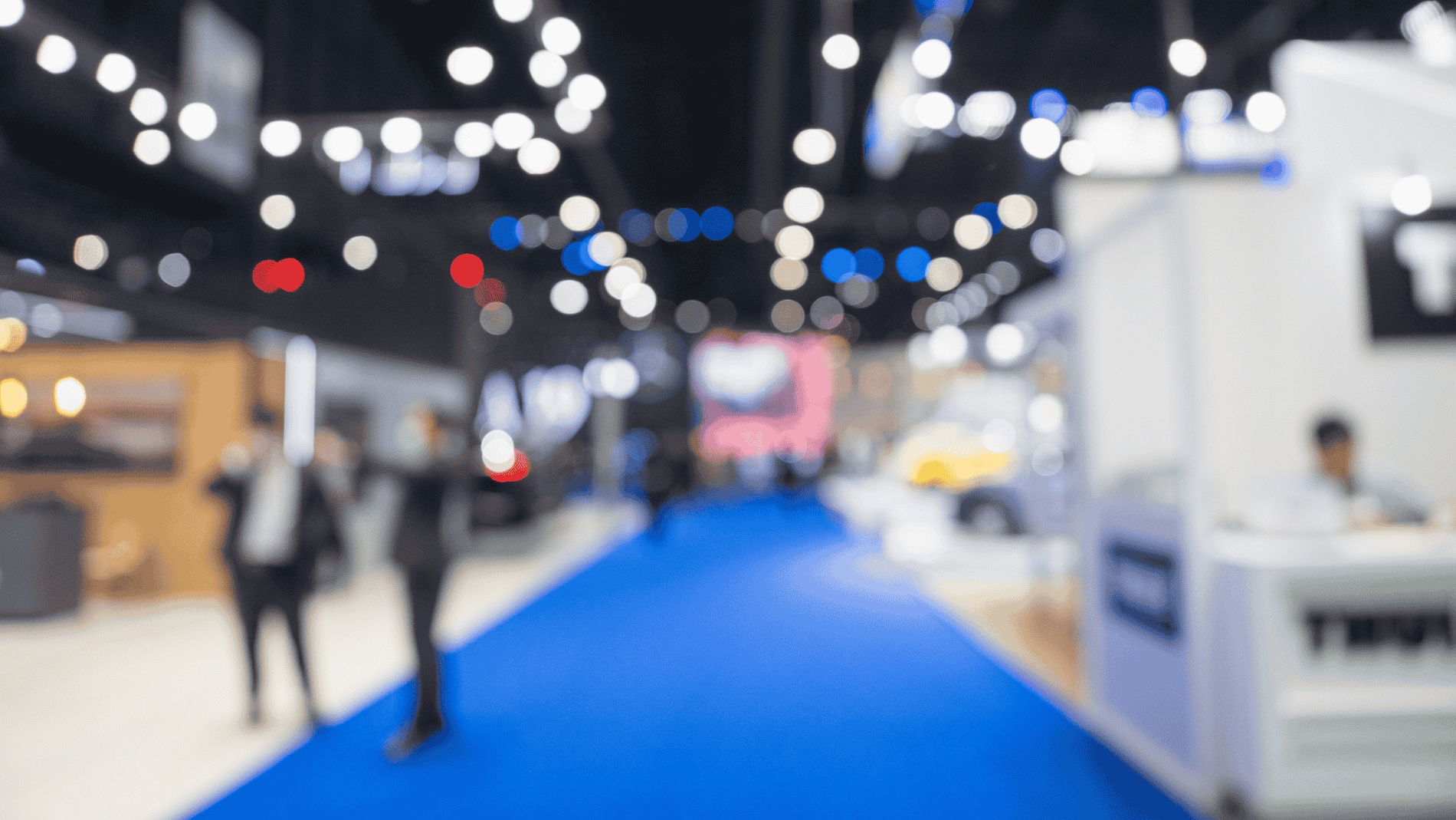 Blurred view of a busy exhibition hall with a blue carpet and various booths.