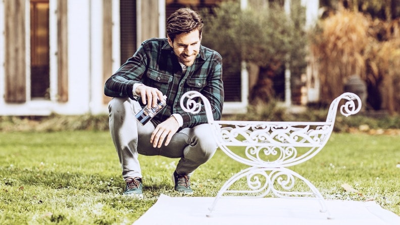 Mann mit einer Sprühdose Metallschutzlack von Hammerite in der Hand hinter einem weißen Metallobjekt in einem Garten.