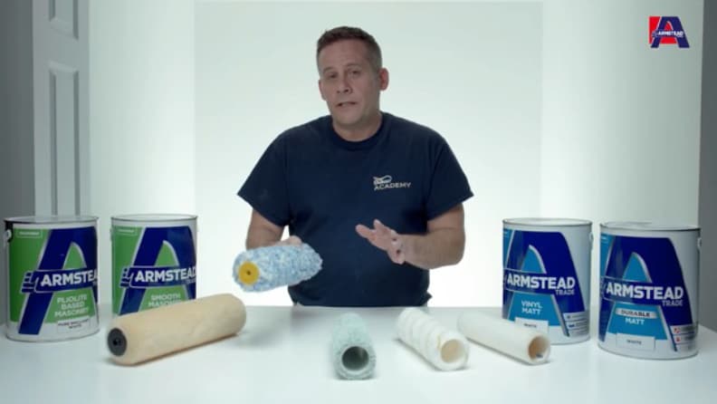 Man in navy blue shirt presenting various Armstead paint products and painting tools including paint cans, roller covers, and brushes on a white table with the Armstead logo visible in the corner.