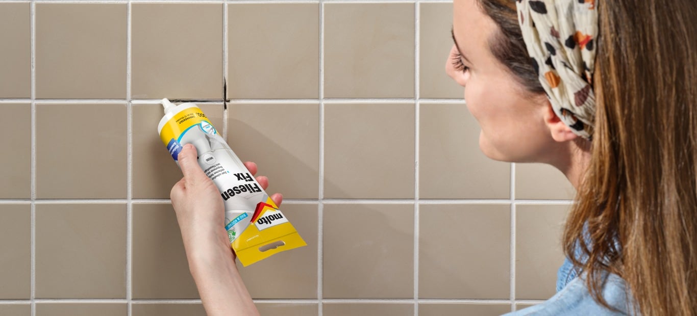 Eine Frau füllt eine herausgebrochene Fuge zwischen Fliesen im Bad mit Fliesen Fix von Molto direkt aus der Tube.
