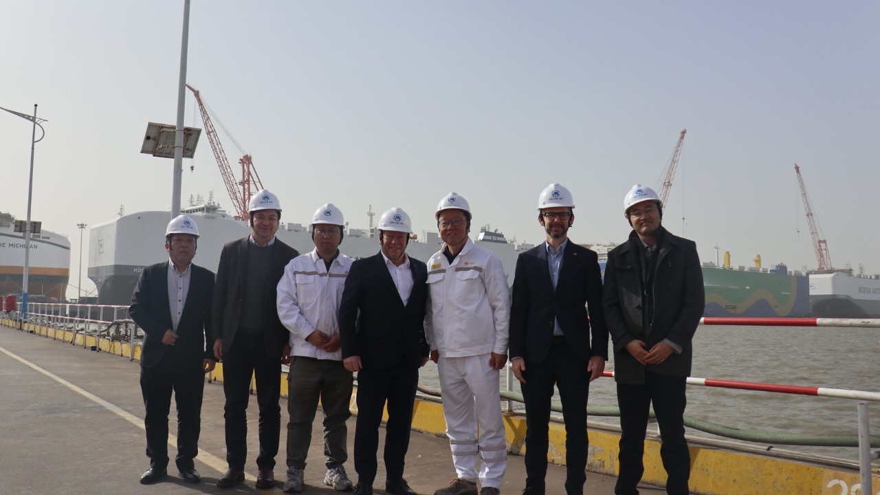 Group of seven men wearing hard hats standing at a dock with ships in the background.