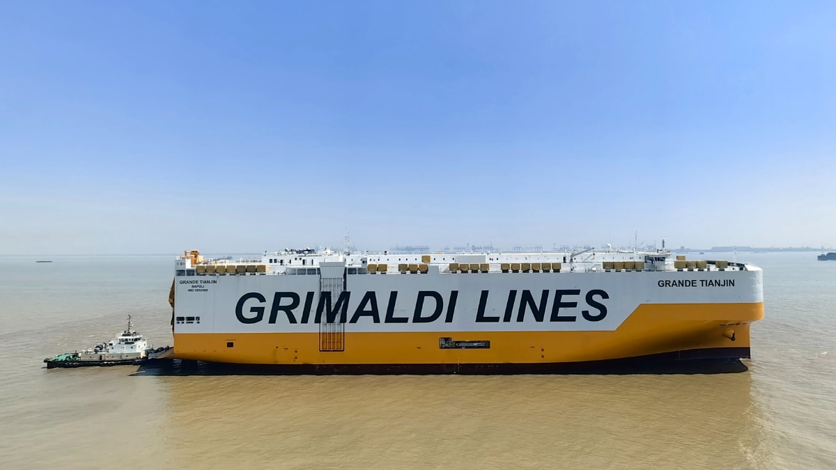 Large vehicle carrier ship branded “GRIMALDI LINES” on the side, sailing through calm brown water under a clear blue sky, with a small tug boat alongside its stern and rows of vehicles visible on the open top deck.