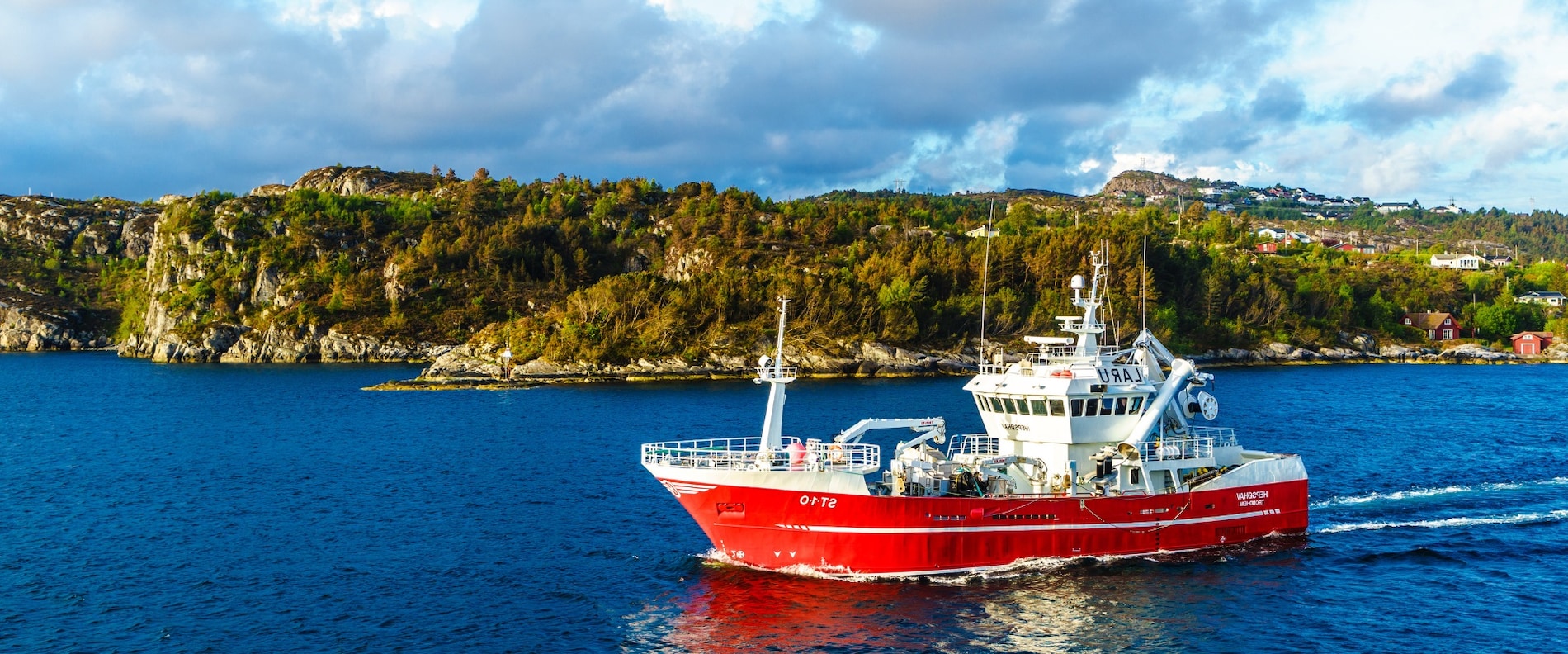 Red fishing vessel sailing in calm coastal waters with rocky shoreline in the background.