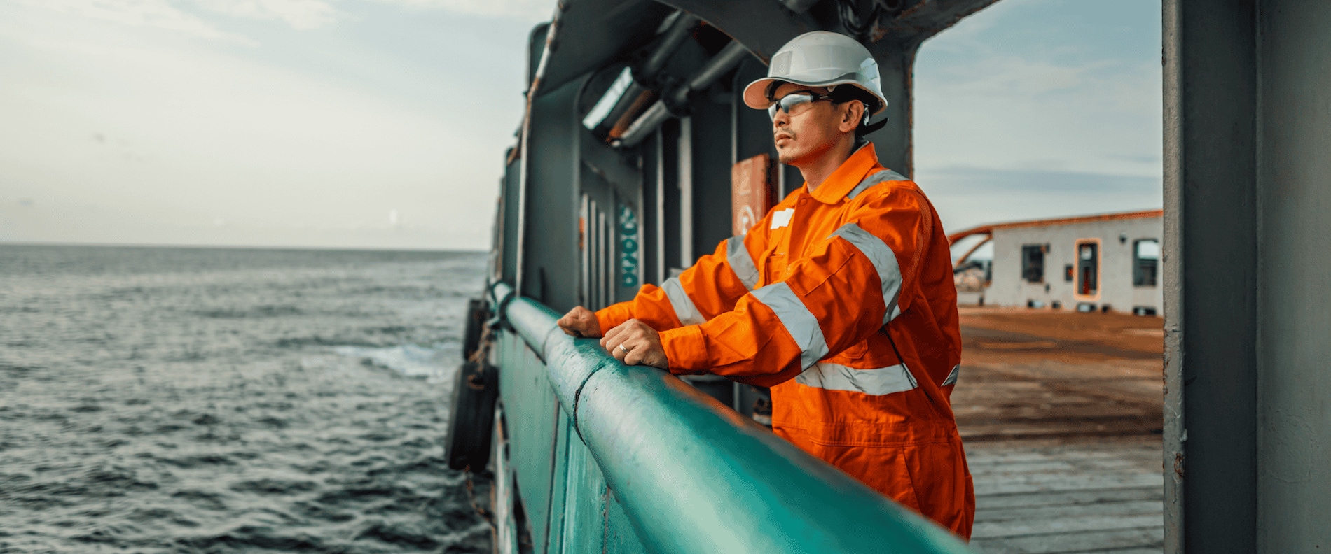 Marine crew member in orange safety gear and helmet standing on a ship deck, looking out over the ocean.