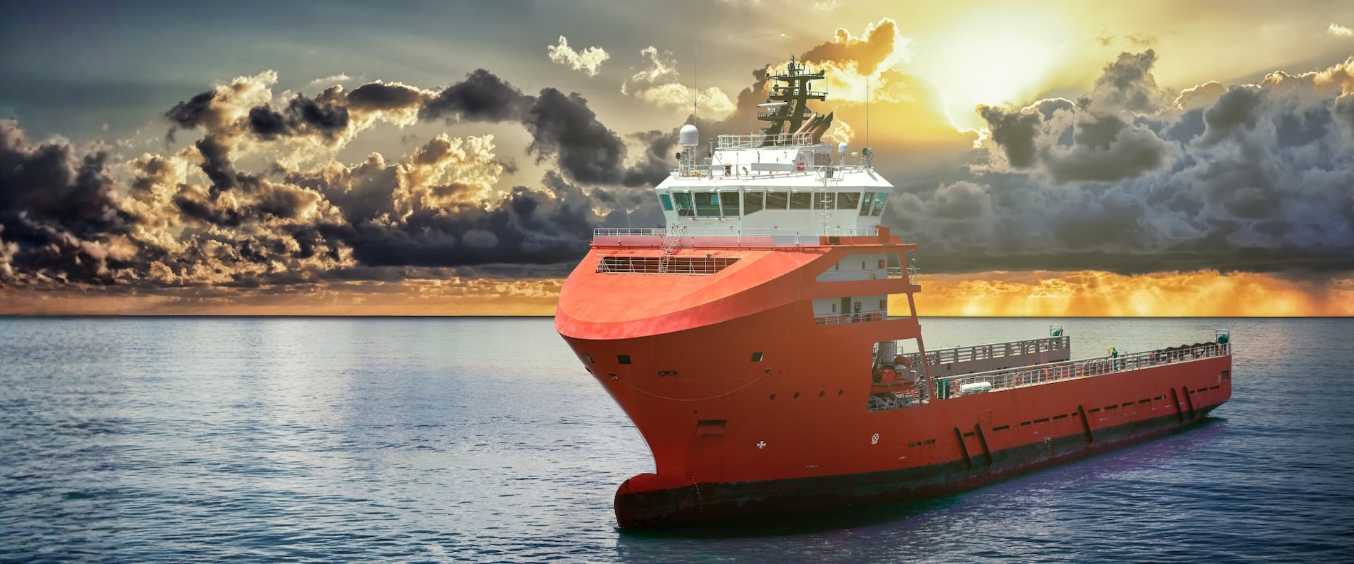 Red offshore support vessel sailing at sea under a cloudy sunset sky.