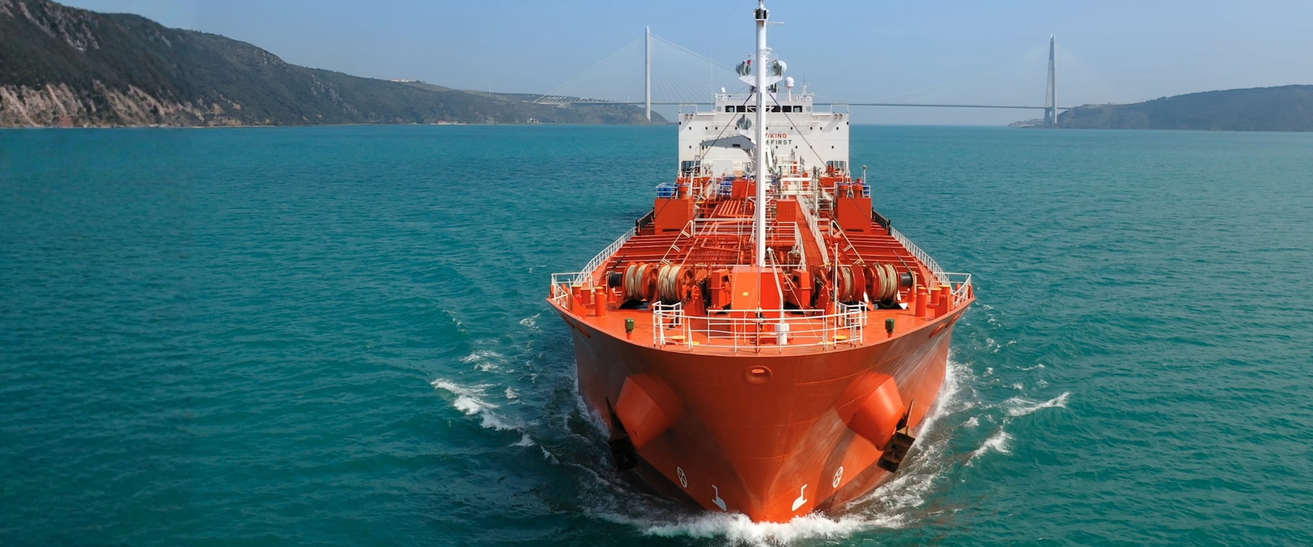 Front view of a red tanker ship sailing through calm blue water with a bridge in the background