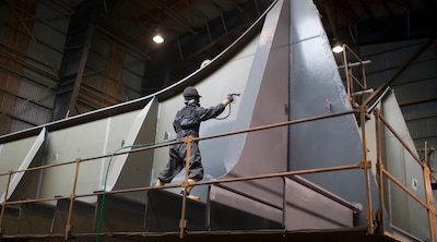 Worker applying protective coating to a steel ship section in a shipyard.