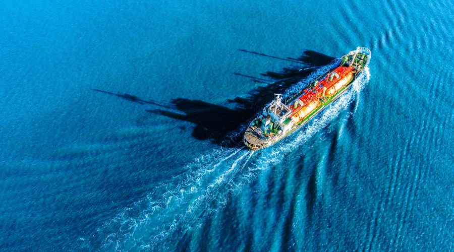 Aerial view of a large commercial vessel sailing through calm blue water, leaving a smooth wake behind it.