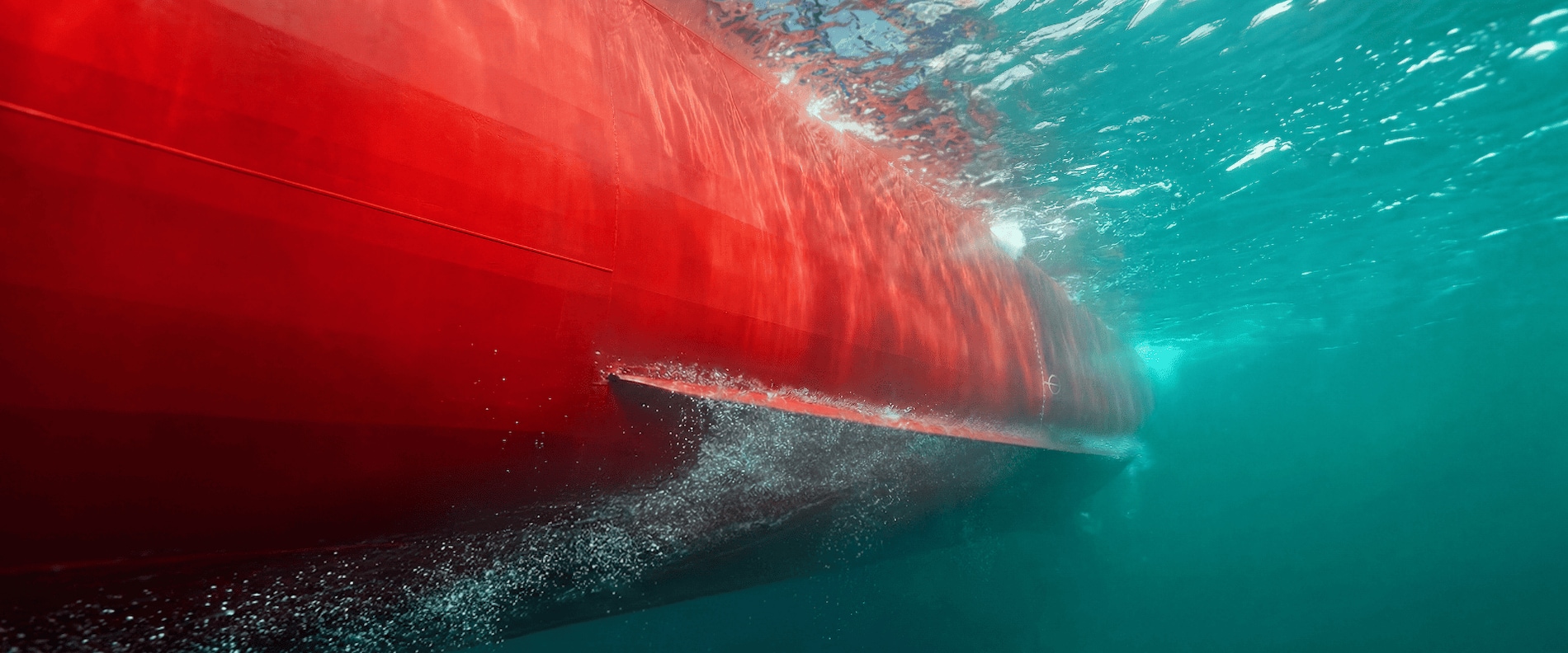 Underwater view of a vessel hull coated with red marine coating moving through clear blue water.