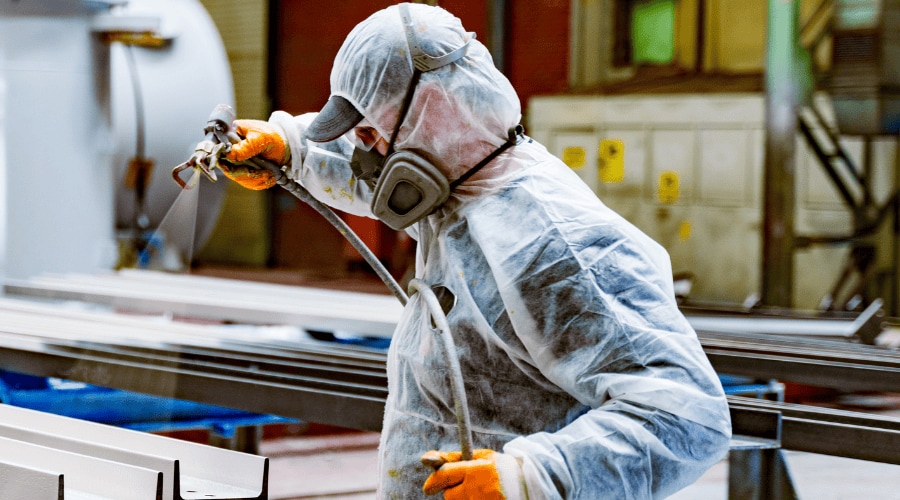 Worker in protective suit applying protective coating to steel framework in manufacturing facility.