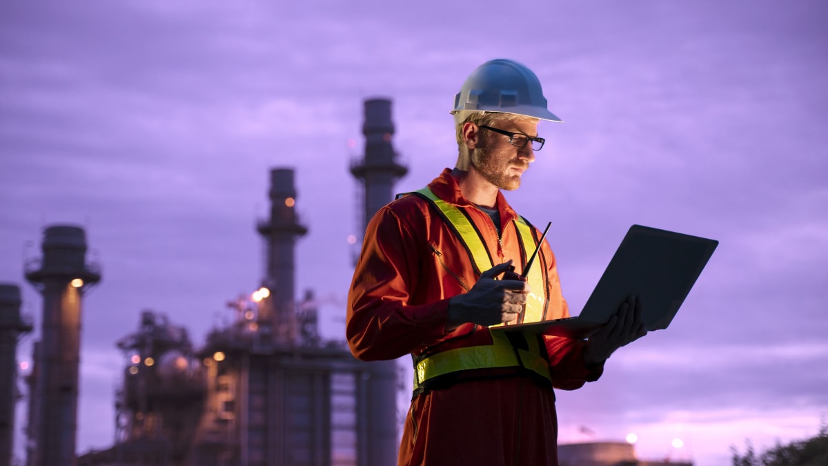 Engineer wearing safety gear uses a laptop while inspecting an industrial refinery at dusk, representing performance monitoring and fire protection technology in energy facilities.
