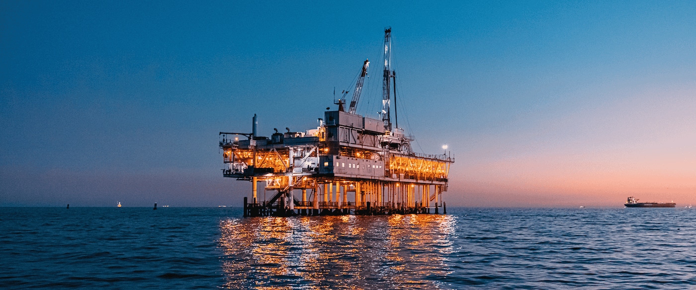 Offshore oil and gas platform illuminated at dusk, standing in calm open water with reflections on the sea surface.