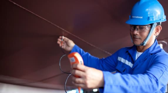 Engineer wearing blue protective gear and helmet inspecting coated steel surface with a digital gauge.
