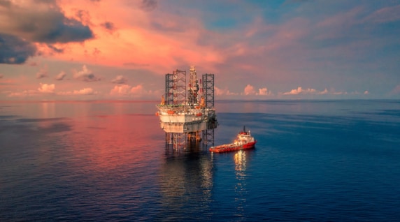 An offshore oil and gas platform standing in calm open water at sunset, with a red support vessel positioned alongside and the sky lit with pink and blue clouds.