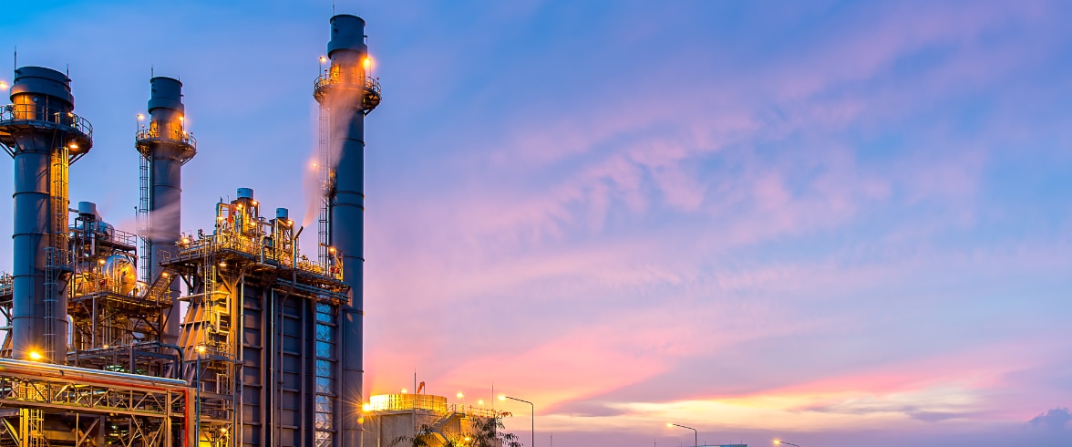 Industrial refinery or processing plant featuring tall metal towers, interconnected pipes and platforms, illuminated in the early evening with a vivid pink and blue sunset in the background.