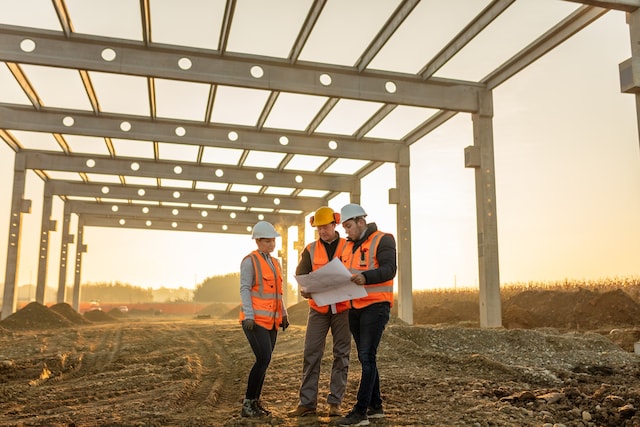 Construction workers wearing safety helmets  and high-visibility vests reviewing plans at a steel frame construction site.