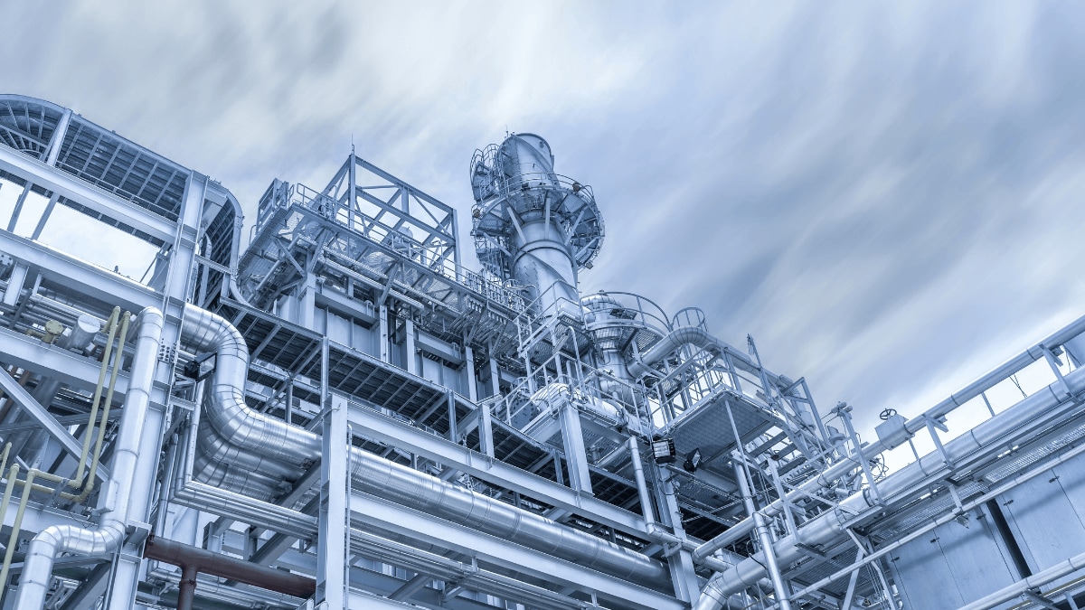Large industrial plant with complex steel piping and a central processing tower beneath a cloudy sky.
