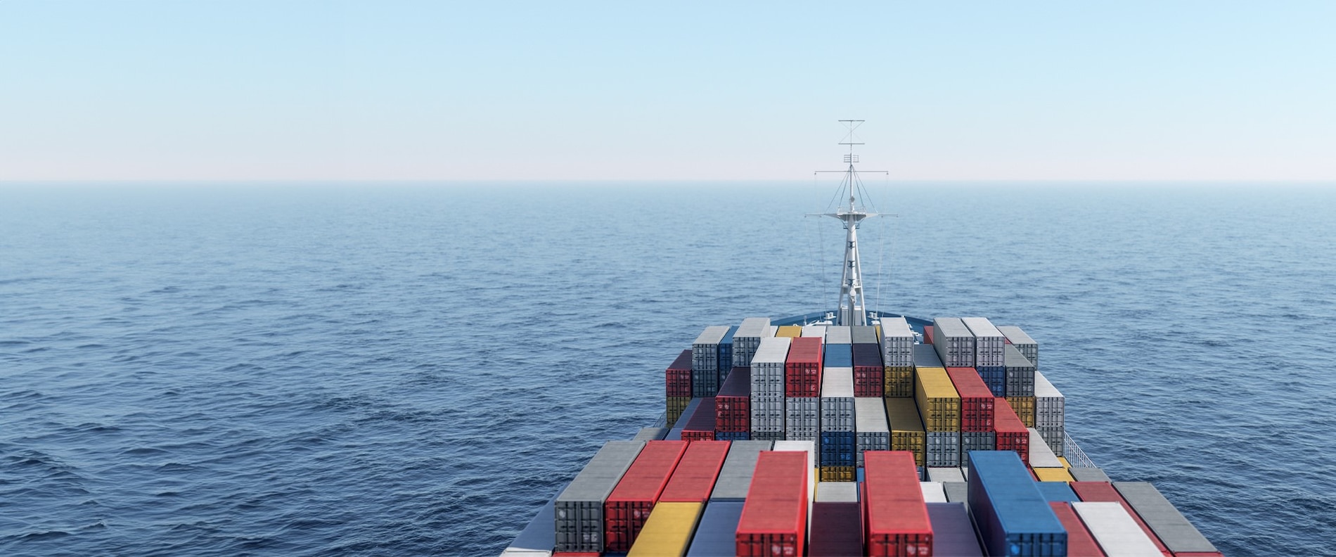 Stacked shipping containers on a large cargo vessel at sea, shown from above the deck, with the ship’s communications mast rising behind the containers, under a wide blue sky that fades into the horizon.