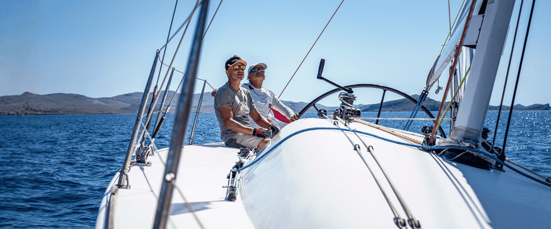 Two people sit near the helm of a sailing yacht, looking ahead as the boat glides across calm blue water with hills in the distance.