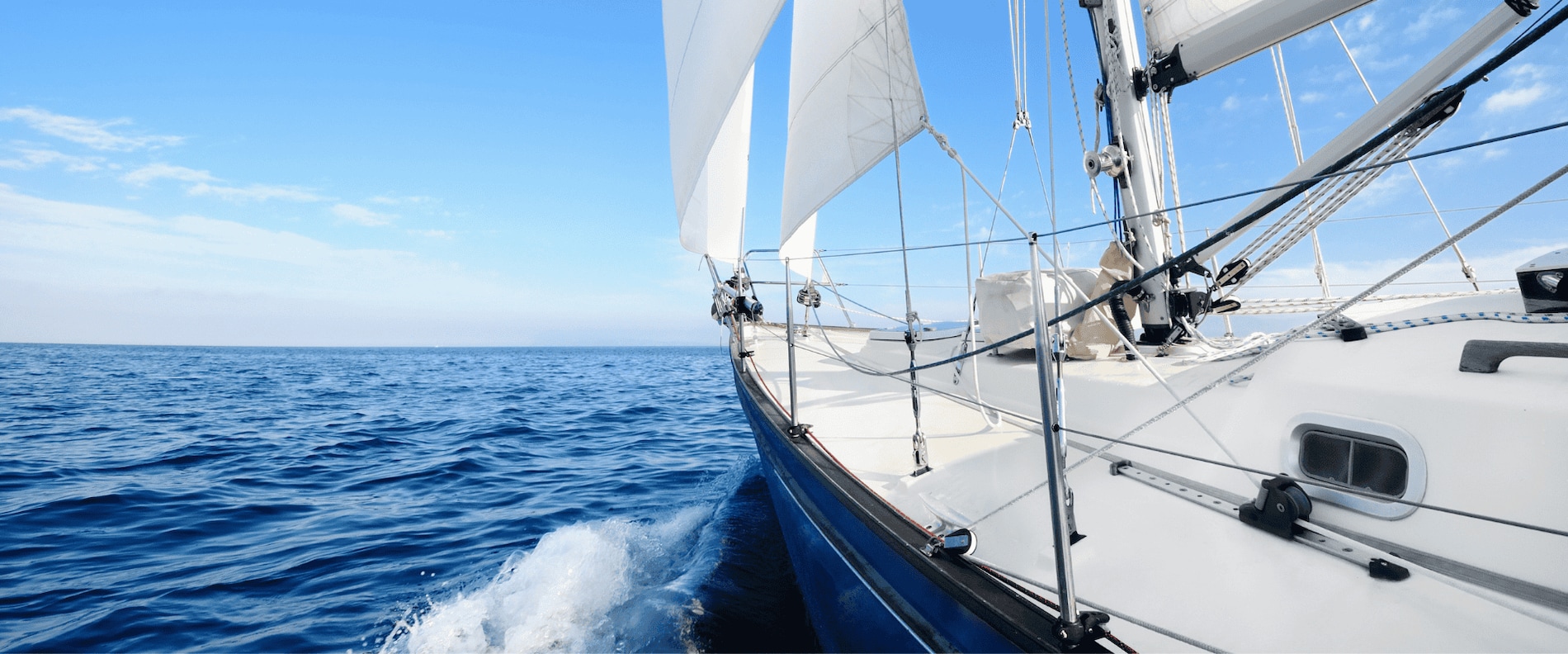 iew from the deck of a sailing yacht moving across calm blue water with white sails raised.