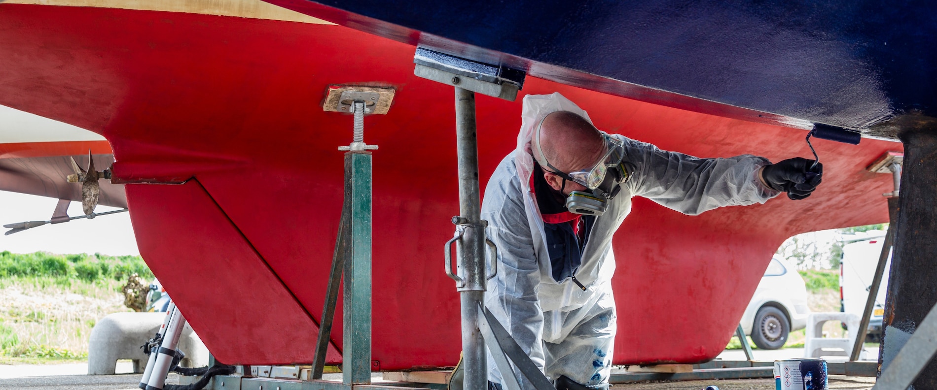 Eine Person in Schutzkleidung trägt eine Beschichtung auf das Unterwasserschiff eines Bootes auf, während sie unter dem Schiff in einer Werft kniet.