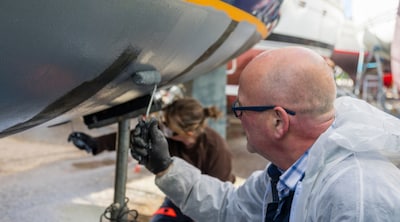 En person i skyddskläder applicerar med en liten färgroller en beläggning på båtens undervattenskrov under underhållsarbete i en varvsmiljö.