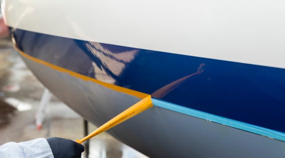 Eine Hand mit Schutzhandschuh entfernt gelbes Abklebeband von der Wasserlinie eines Segelbootrumpfs während der Vorbereitung des Unterwasserschiffs.