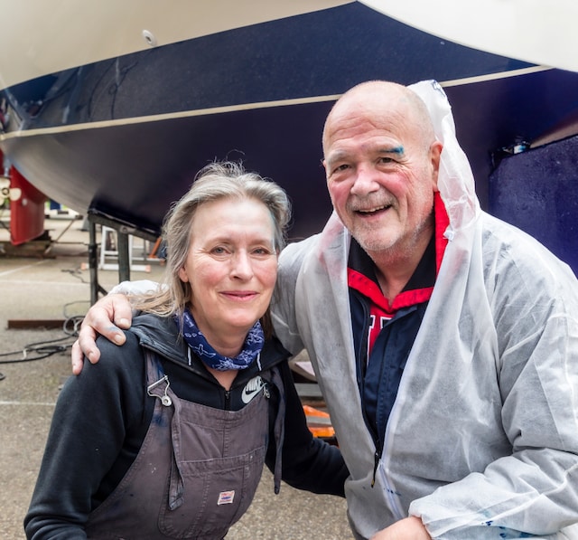 Zwei Personen in Arbeitskleidung stehen lächelnd zusammen in einer Werft vor einem Segelbootrumpf nach Abschluss von Wartungsarbeiten.