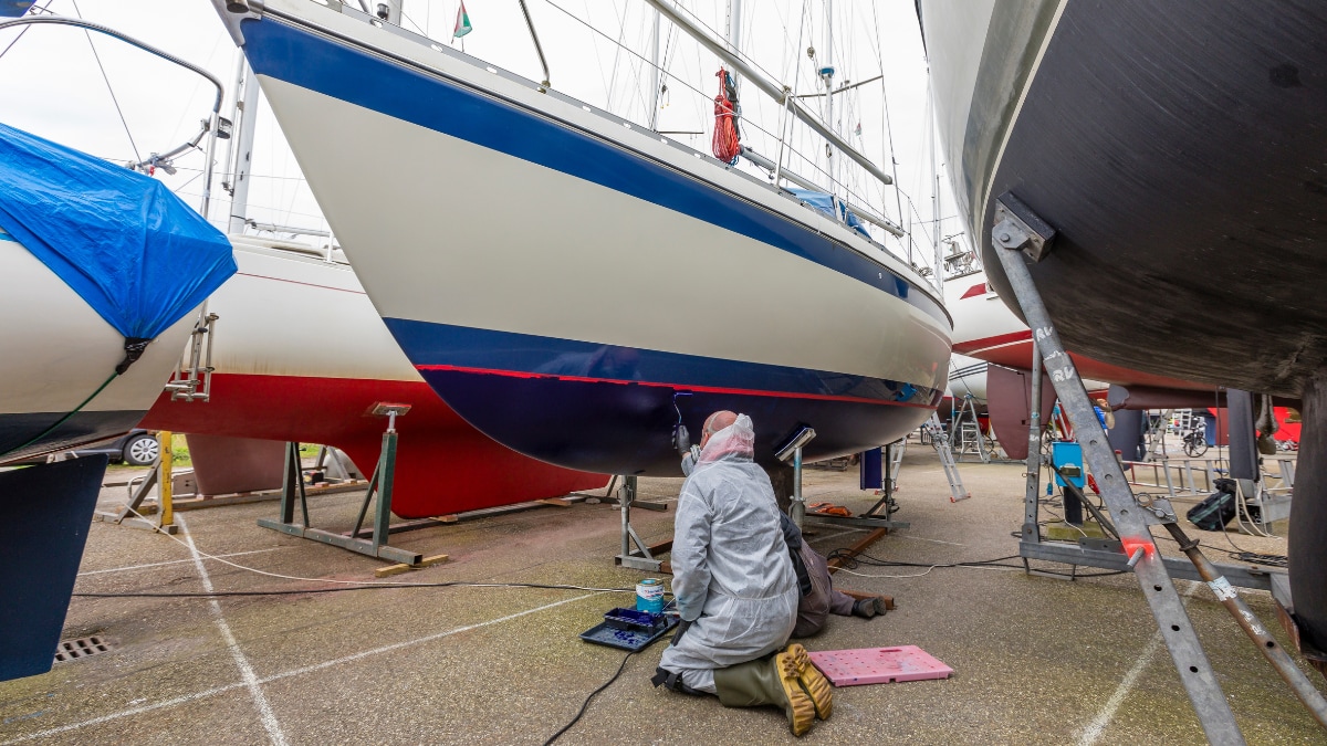 Eine Person in Schutzkleidung kniet in einer Werft unter einem Segelboot und trägt eine Beschichtung auf das Unterwasserschiff auf, während weitere Boote auf Böcken in der Umgebung stehen.