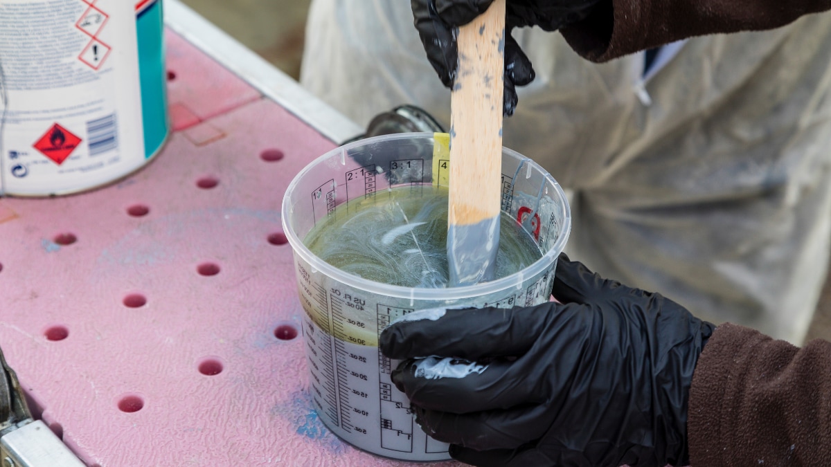 Mit Handschuhen geschützte Hände rühren eine Beschichtung in einem transparenten Mischbecher an.