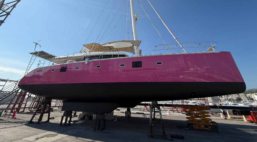A 62ft Lagoon catamaran with a pink hull and black underwater coating standing on supports in a boatyard after the restoration.