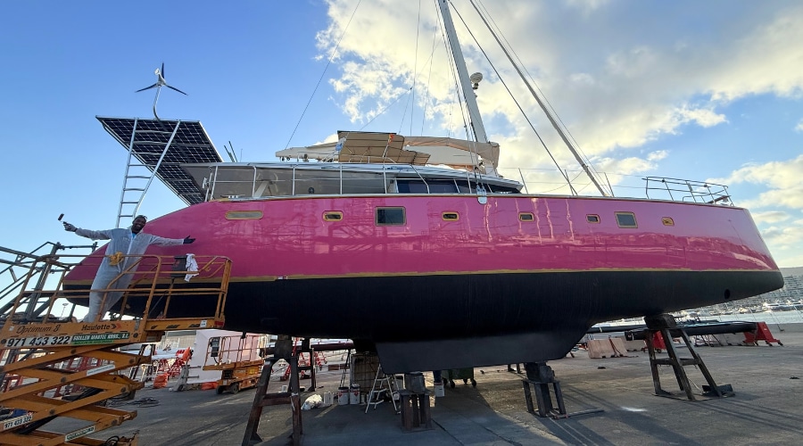 A 62ft Lagoon catamaran with a pink hull and black antifouling standing on supports in a boatyard in Mallorca during the refit.
