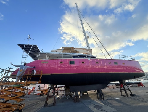 A 62ft Lagoon catamaran with a pink hull and black antifouling, supported on stands in a boatyard in Mallorca during the refit.