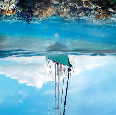 En undervattensbild av ett segelbåtsskrov med köl och tydlig vattenlinje, medan ett korallrev breder ut sig i klart blått vatten.