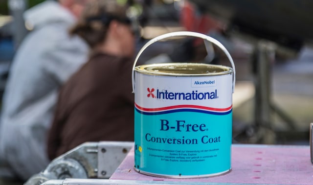 En burk International® B-Free Conversion Coat på en metallplattform i en varvsmiljö, med två personer som arbetar på ett segelbåts undervattenskrov i bakgrunden.
