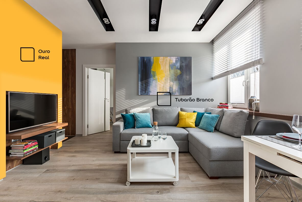 Sala de TV renovada com as cores Tubarão branco e Ouro Real da Tintas Coral. O amarelo super combinou com a tonalidade cinza da parede e também com a decoração em branco da mesinha de centro.