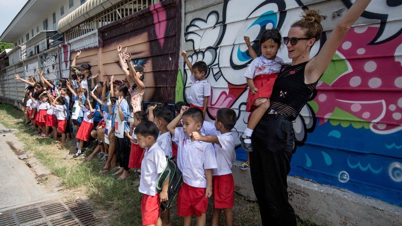 Grupo de crianças e uma mulher frente a uma parede pintada na Tailândia.