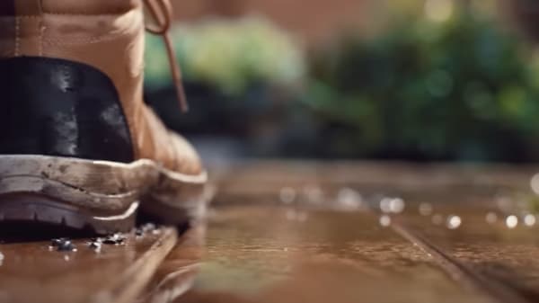 Közeli kép egy barna munkabakancsról nedves, közepesen barna fa teraszon, ahol vízcseppek gyöngyöznek a felületen, vízlepergető védelmet demonstrálva