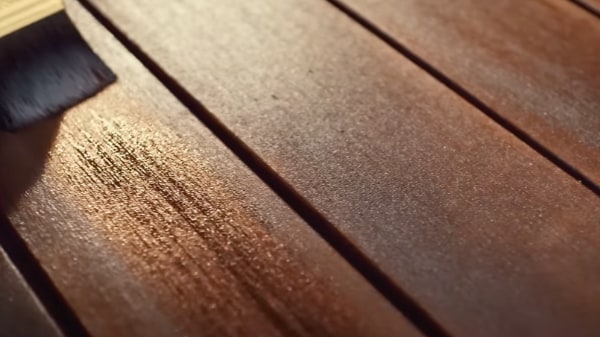 Közeli kép sötét sörtéjű ecsetről, amely gazdag vörösesbarna falazúrt visz fel fadeszkákra, bemutatva a fényes kezelt sötét fa és a világosabb kezeletlen deszkák közötti kontrasztot