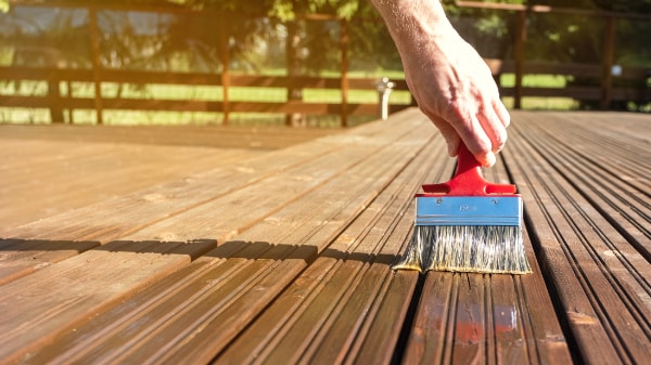 Személy sötét falazúrt visz fel kültéri teraszdeszkára széles ecsettel, amely piros nyelű és kék virolla, bemutatva a gazdag sötét kezelt deszkák és a világosabb kezeletlen fa közötti kontrasztot