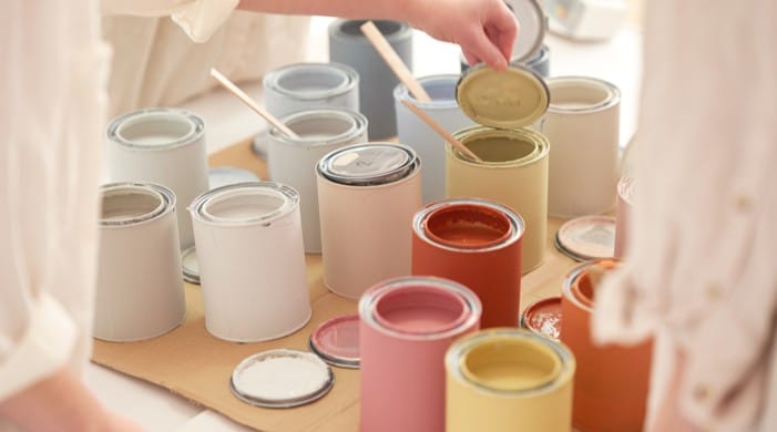                       A tabletop arrangement of open paint tins in soft neutral and warm tones, including white, grey, pink, yellow, and terracotta, with wooden stir sticks inside several cans as a hand lifts the lid from a pale yellow paint tin.
