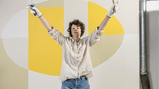 O femeie îmbrăcată în cămașă și blugi stă în fața unui perete decorat cu forme geometrice, vopsit cu lavabilă savana.