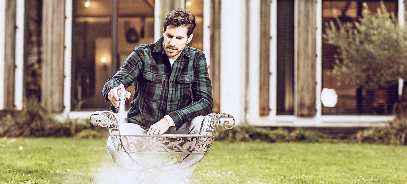 Ein Mann sprüht in einem Garten Metallschutzlack von Hammerite aus der Dose auf ein Metallobjekt