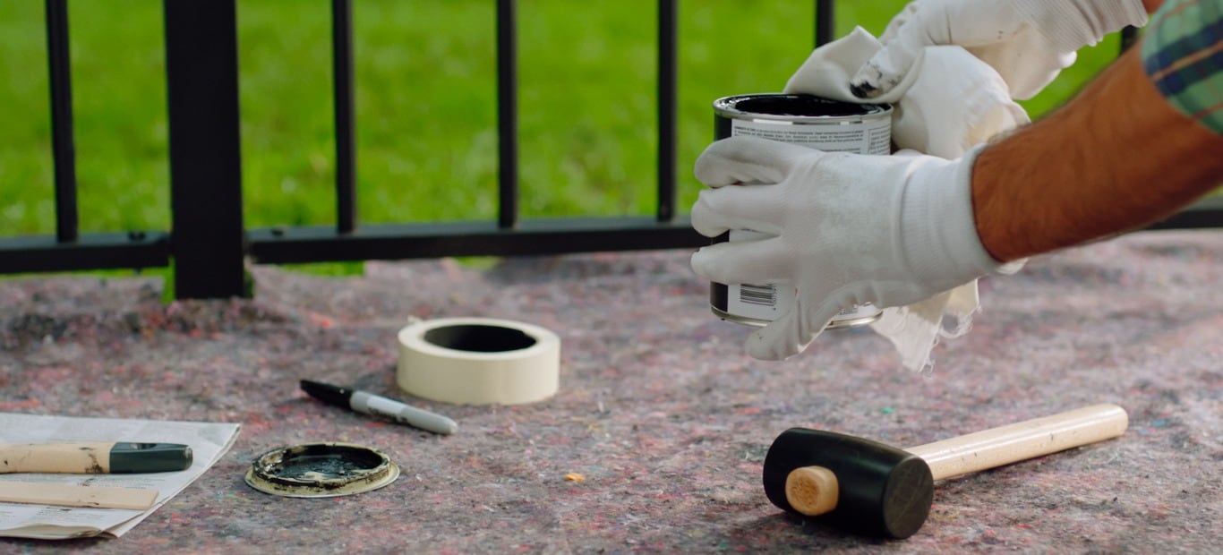 Ein Mann wischt mit einem Tuch Lackreste von einer Dose, um sie luftdicht verschließen und richtig lagern zu können. Auf dem Malervlies darunter liegen ein Gummihammer, eine Rolle Malerkrepp, der Deckel der Dose und Pinsel.