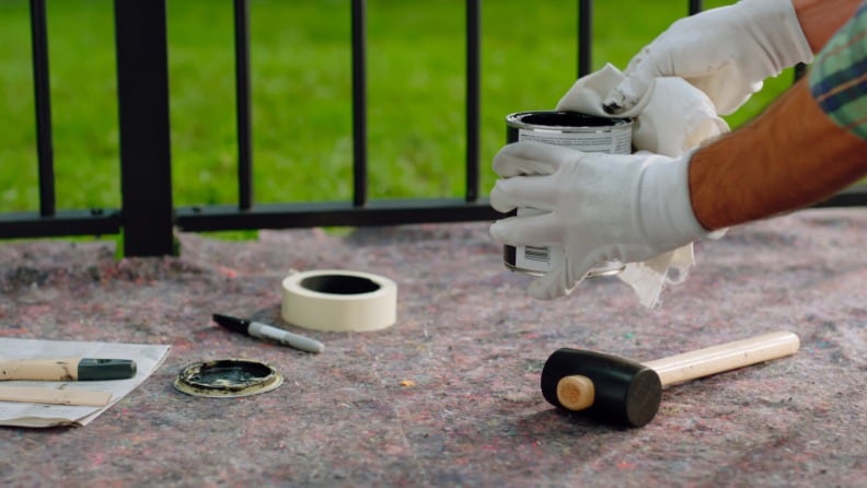 Ein Mann wischt mit einem Tuch Lackreste von einer Dose, um sie luftdicht verschließen und richtig lagern zu können. Auf dem Malervlies darunter liegen ein Gummihammer, eine Rolle Malerkrepp, der Deckel der Dose und Pinsel.
