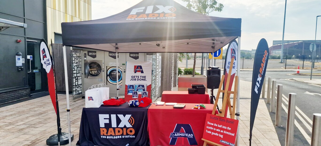 Outdoor promotional tent setup featuring Armstead and Fix Radio branding with red display tables, promotional materials, banners, and a basketball hoop game in an urban plaza setting.